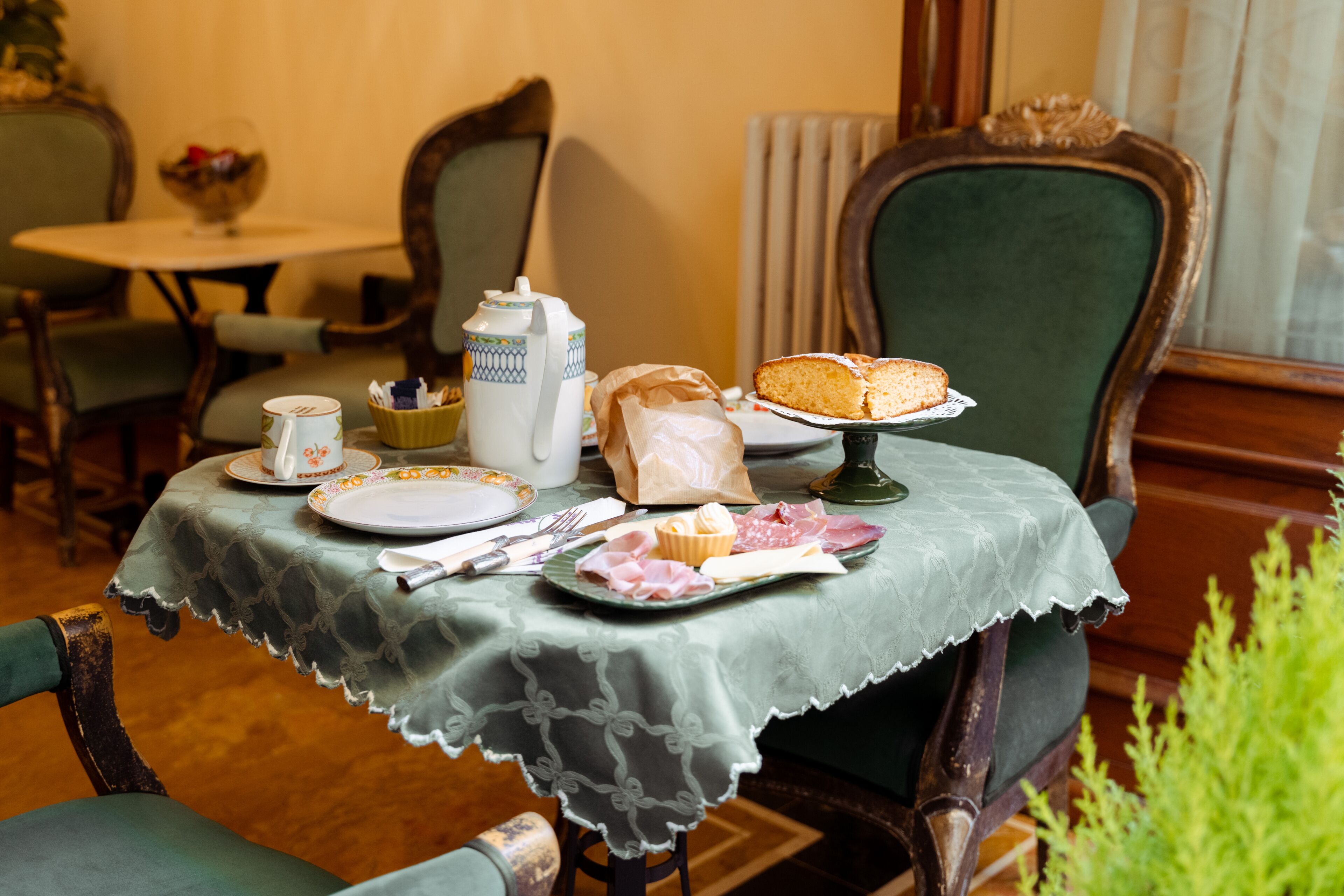 A dining table with a lace tablecloth, plates, a teapot, a cake, and a bag of snacks.