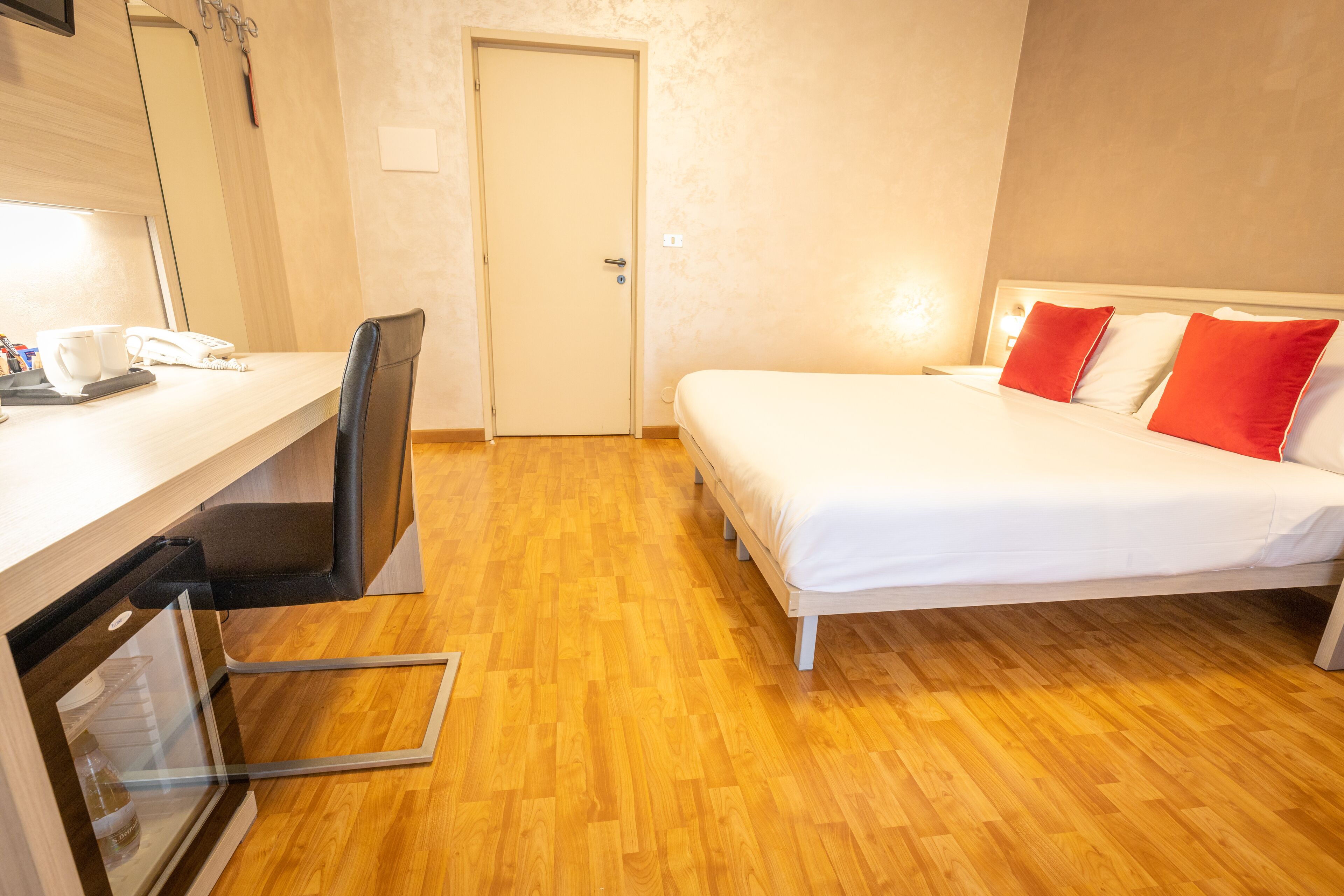 Une chambre d’hôtel avec un plancher en bois, un lit avec des oreillers blancs et rouges, un bureau avec un téléphone et des tasses, et une porte.