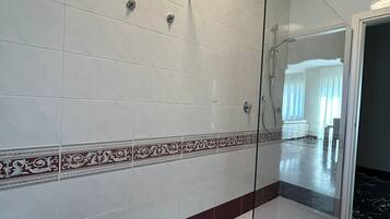 Un baño moderno con cabina de ducha de vidrio, azulejos blancos y granate, y un área de baño visible al fondo.