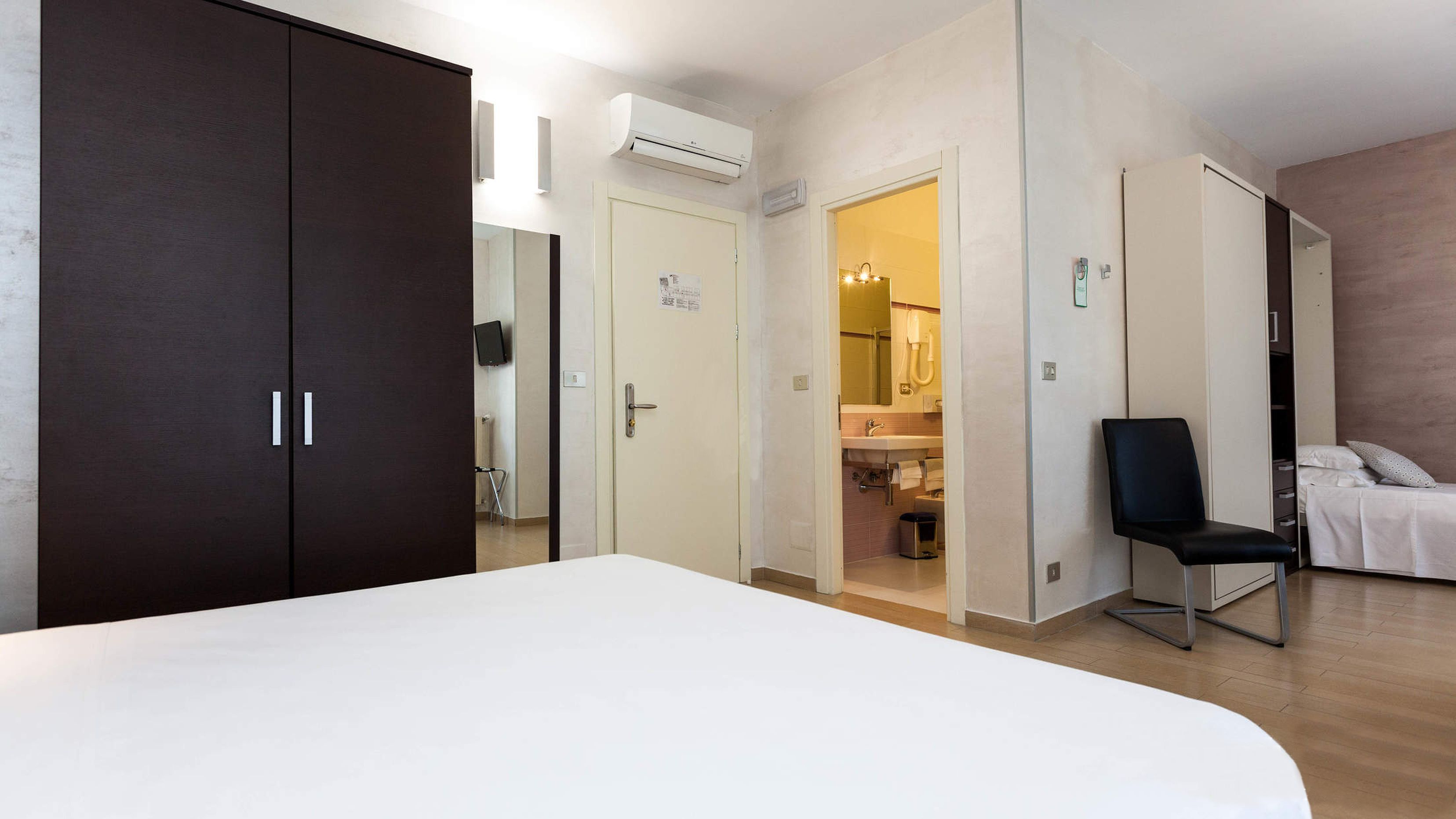 Une chambre à coucher avec un grand lit, une armoire, une chaise et une salle de bain visible à travers une porte ouverte.