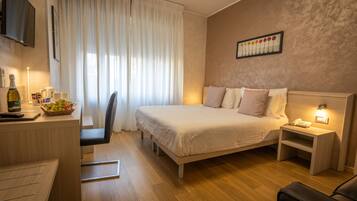 A hotel room with a bed, bedside table with a lamp, a desk with a bottle, and a chair.