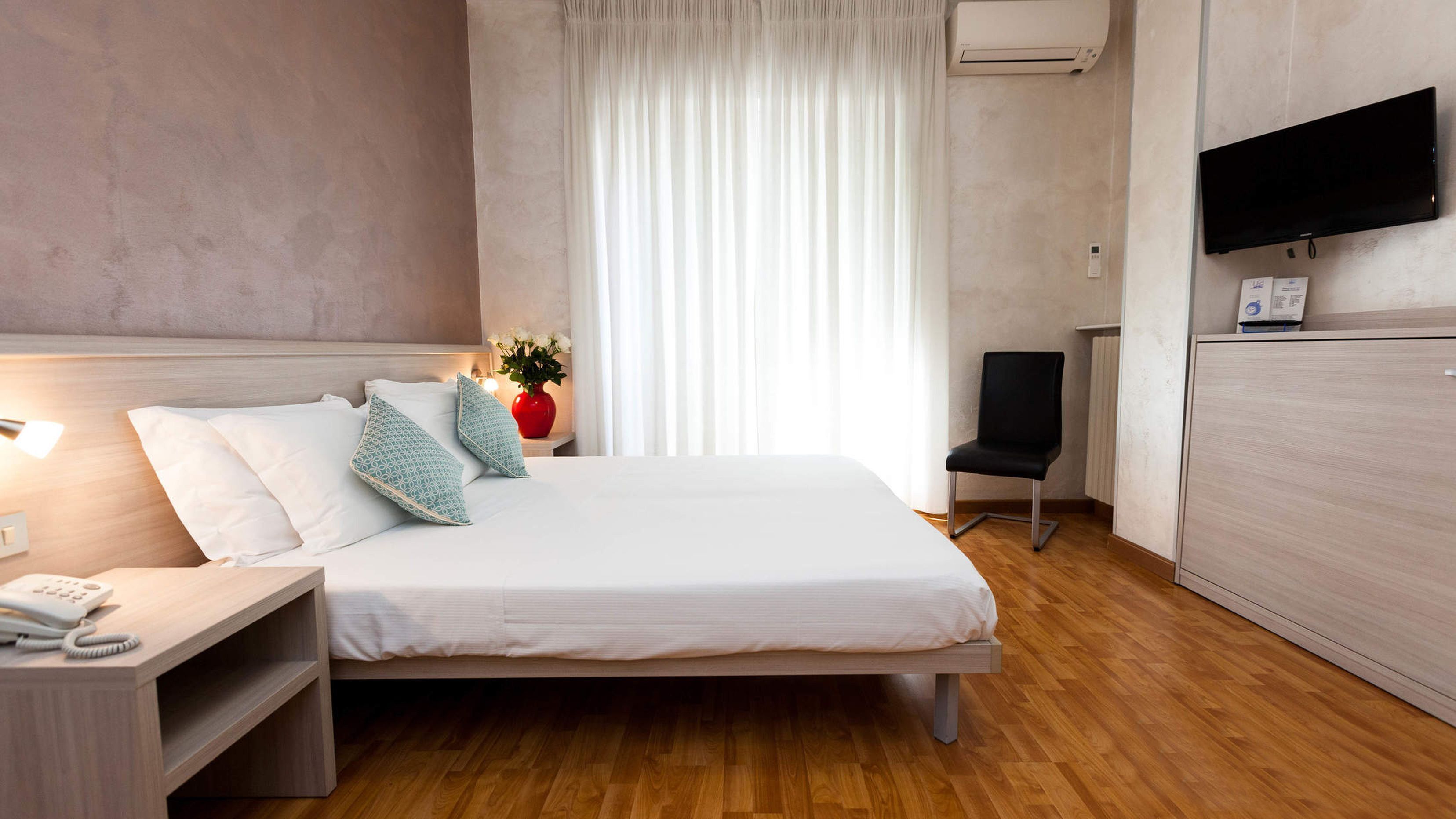 Una habitación de hotel moderna con una cama grande, una mesita de noche con un teléfono, una silla, un televisor montado en la pared y una ventana con cortinas.