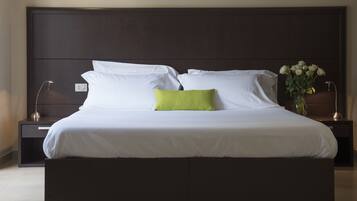 Una cama bien hecha con almohadas blancas y verdes, un cabecero oscuro y un jarrón con flores blancas sobre una mesita de noche.