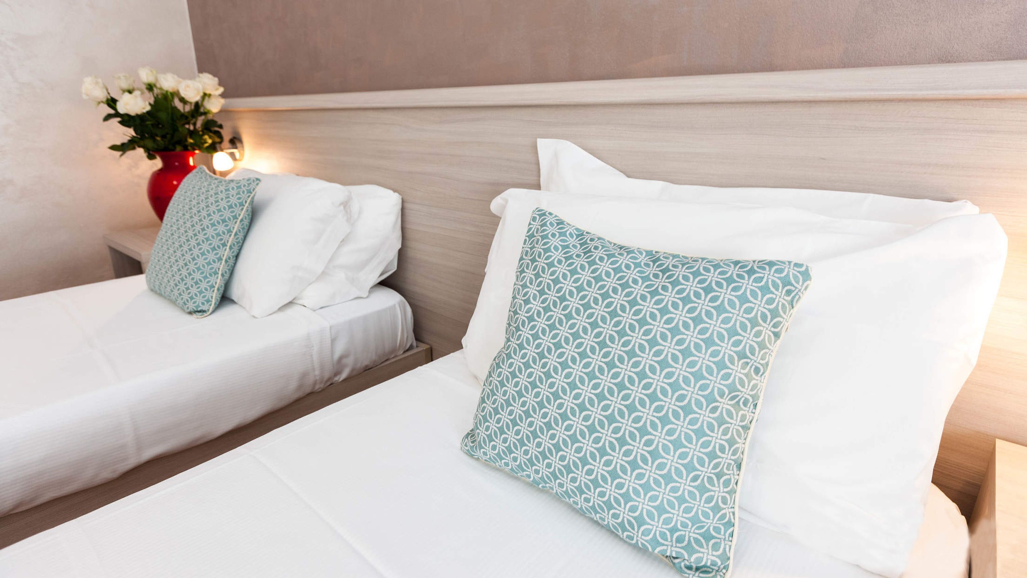 A hotel room with two beds, each with white linens and patterned pillows. A vase with white flowers is on the nightstand.
