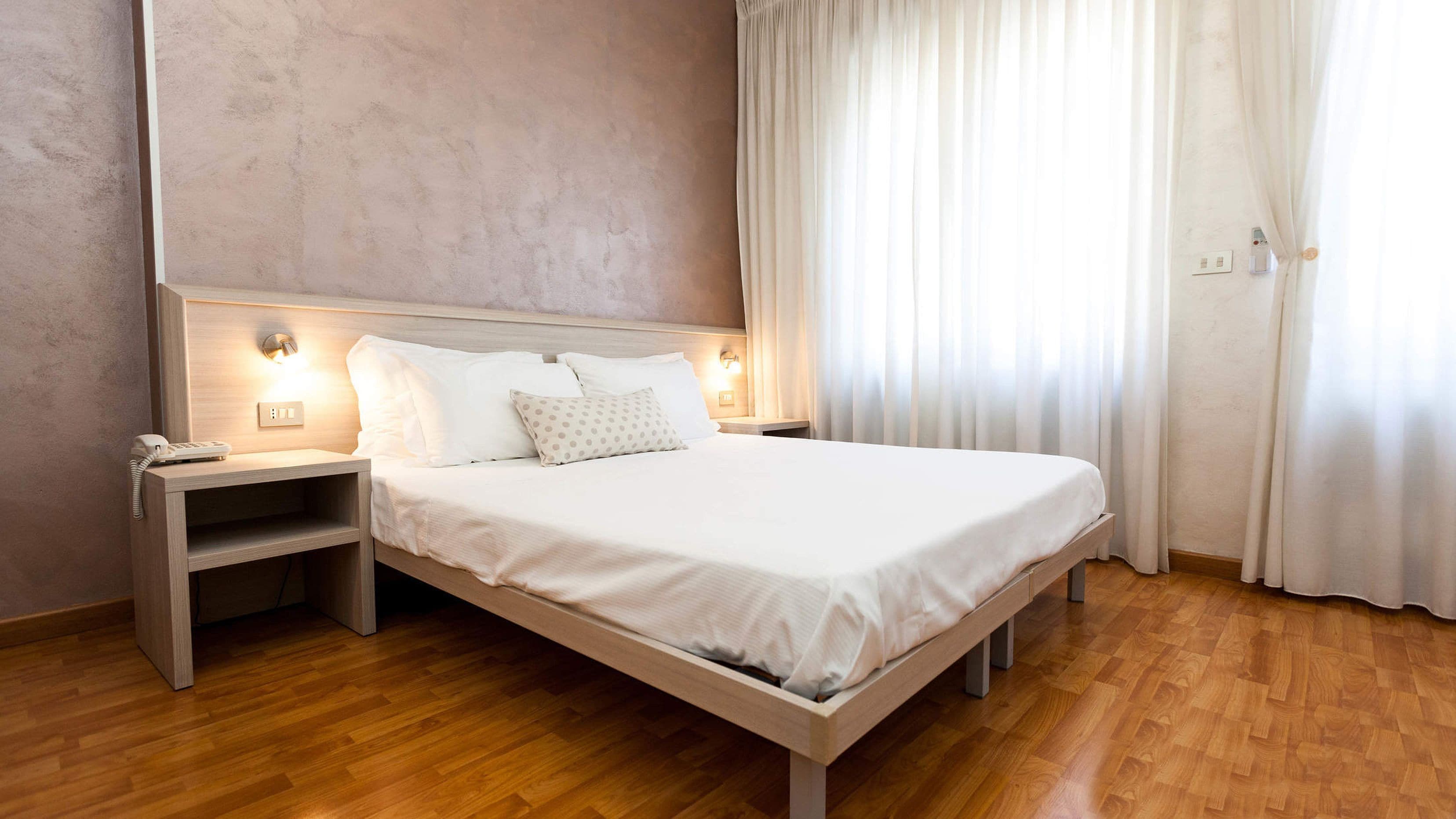 Ein ordentlich bezogenes Bett mit weißen Leinen und einem Holzkopfstück, ein Nachttisch mit Telefon sowie eine wandmontierte Lampe.