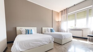 Een moderne hotelkamer met twee bedden, een groot raam met zonwering, een vloerlamp en een radiator.