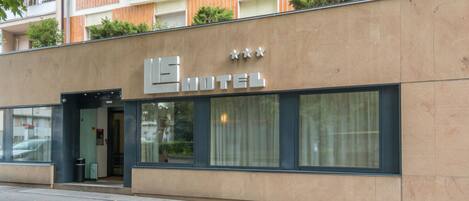 A modern hotel facade with a prominent sign, large windows, and a balcony with greenery.