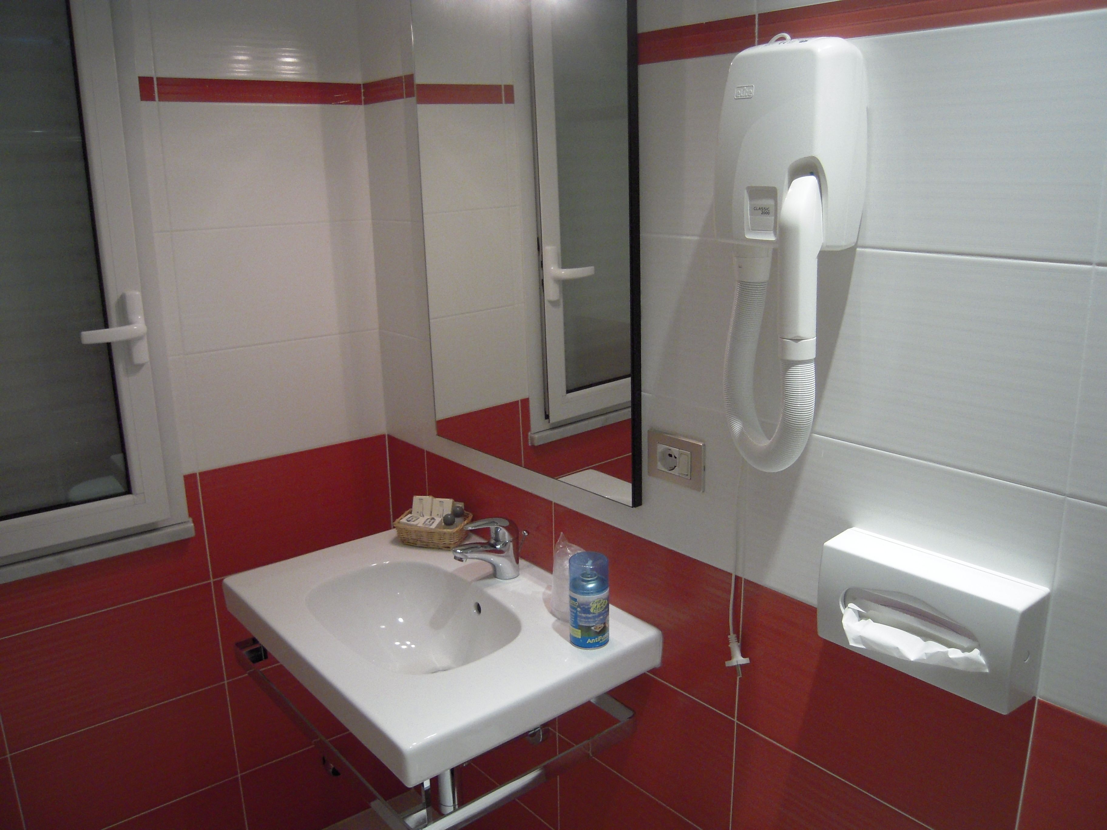 A bathroom with a white sink, red-tiled walls, a mirror, a hand dryer, and a paper towel dispenser.