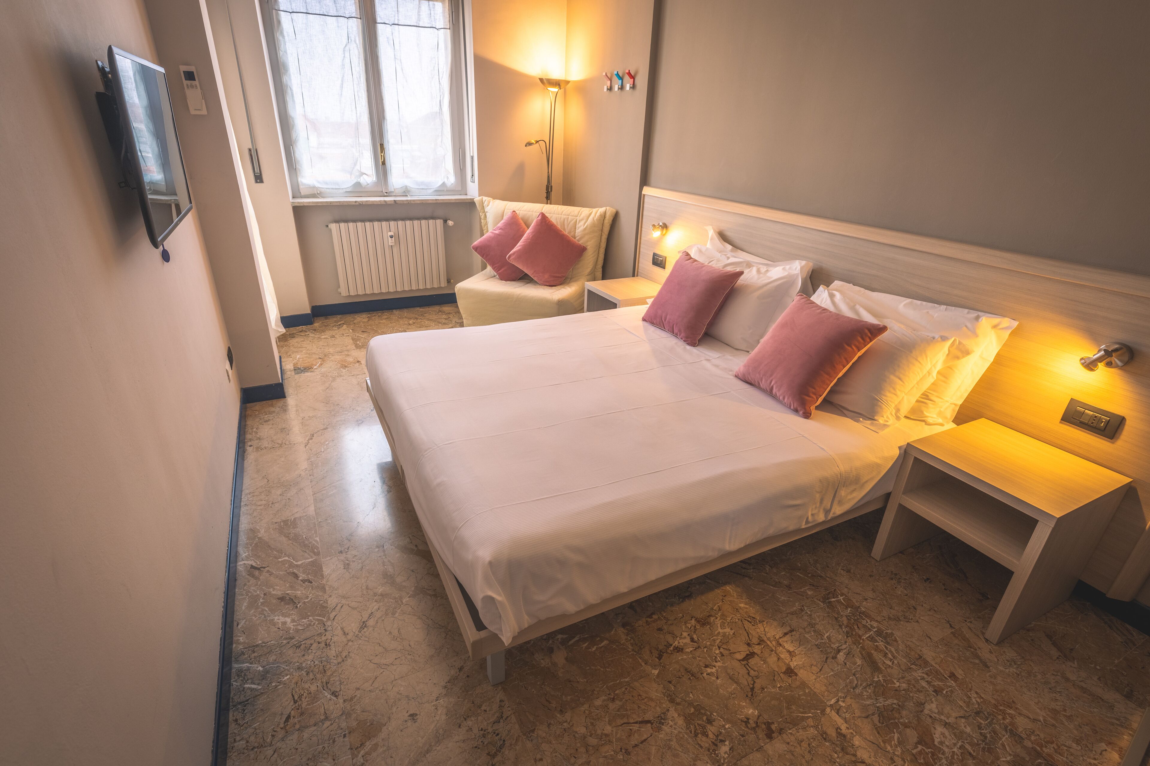 A hotel room with a large bed, bedside tables, a lamp, a TV, and a window with curtains.