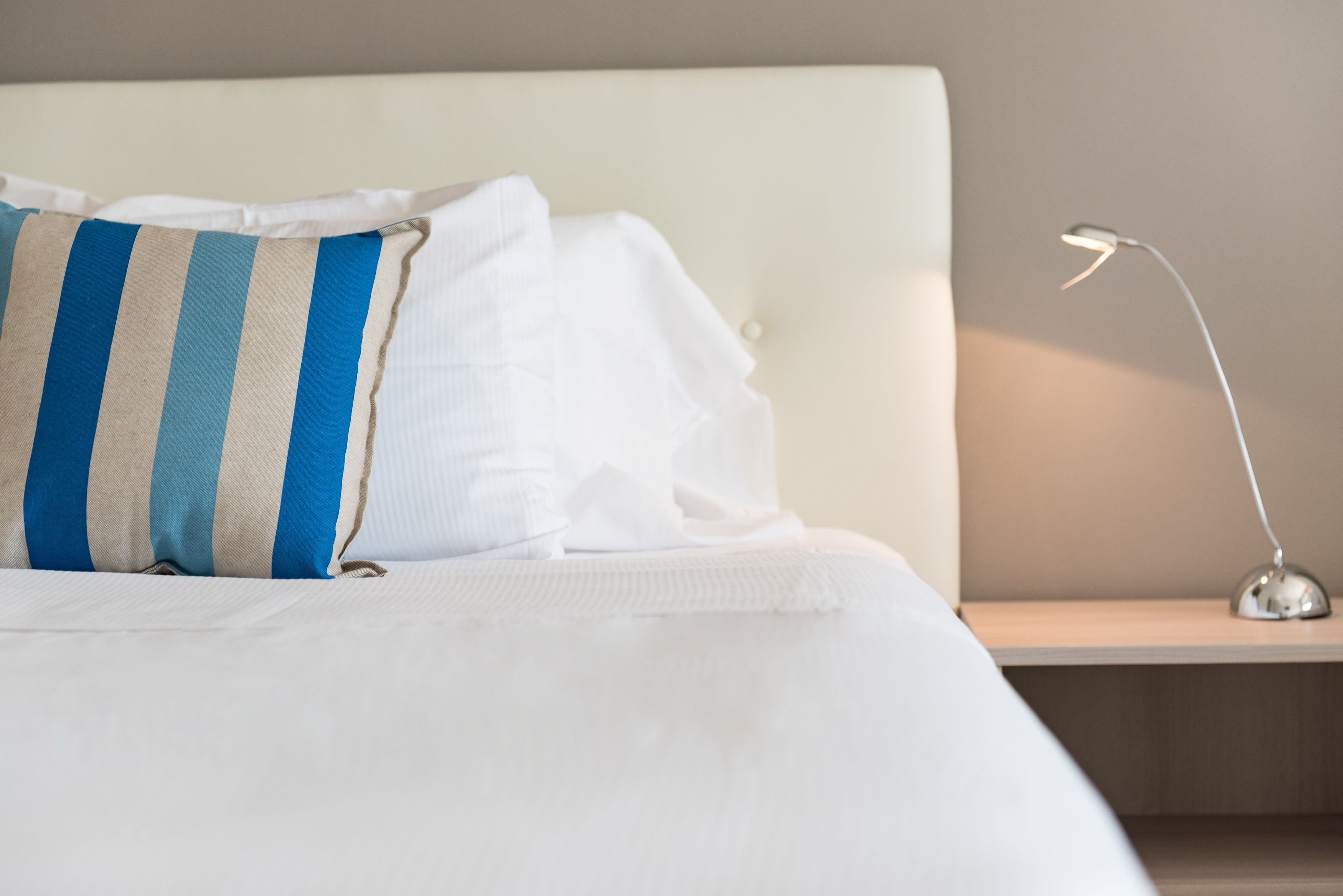 A neatly made bed with white bedding and a striped pillow, a bedside table with a lamp, and a plain wall in the background.