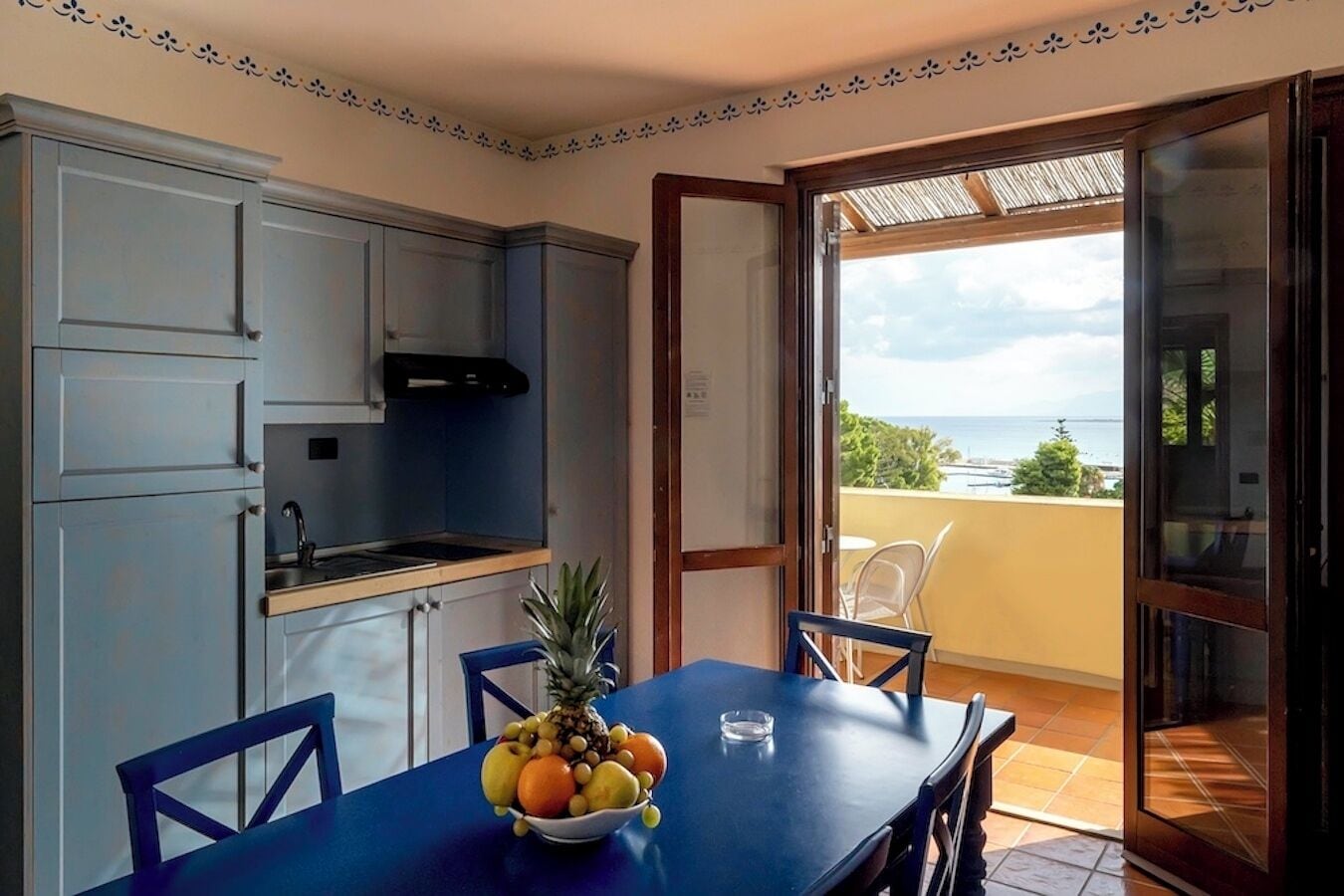 Una cocina con armarios azules, una mesa de comedor con frutero y vista al mar por una puerta abierta.