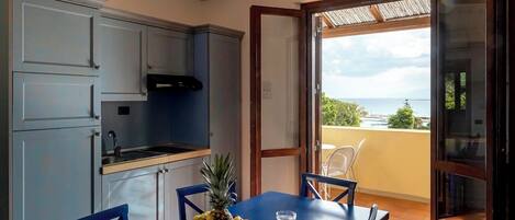 Una cocina con armarios azules, una mesa de comedor con frutero y vista al mar por una puerta abierta.