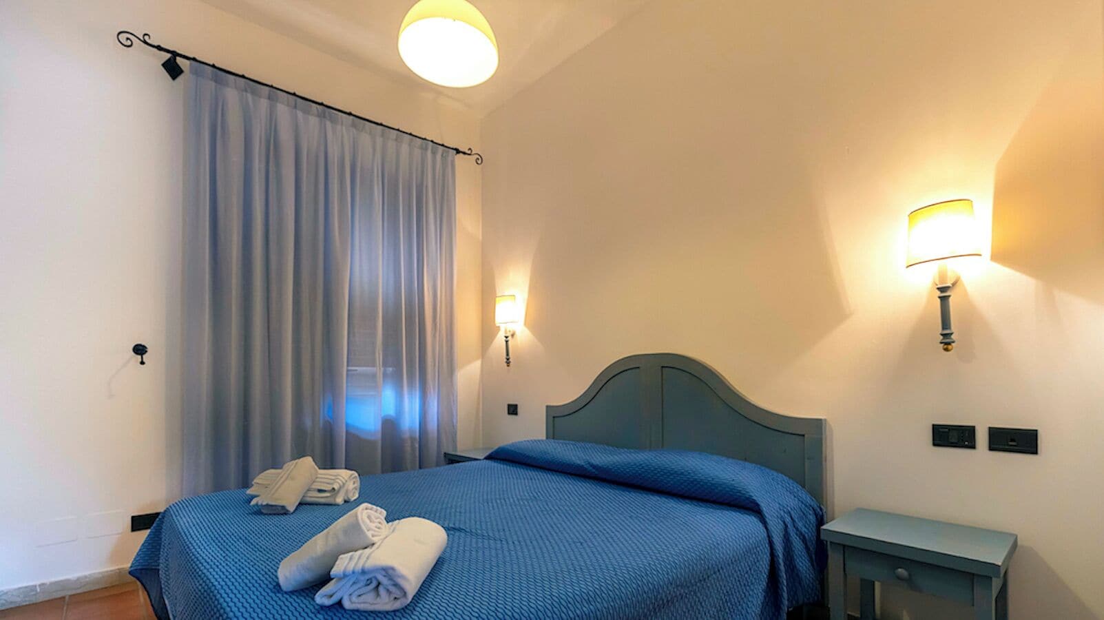 Una habitación de hotel con cama, mesita de noche, lámpara de pared y ventana con cortinas.