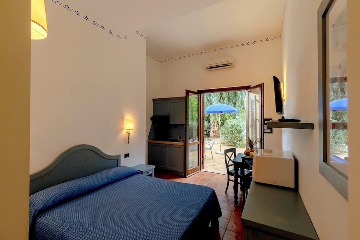 A bedroom with a bed, a desk, and a chair. There is a sliding door leading to an outdoor area with a table and an umbrella.