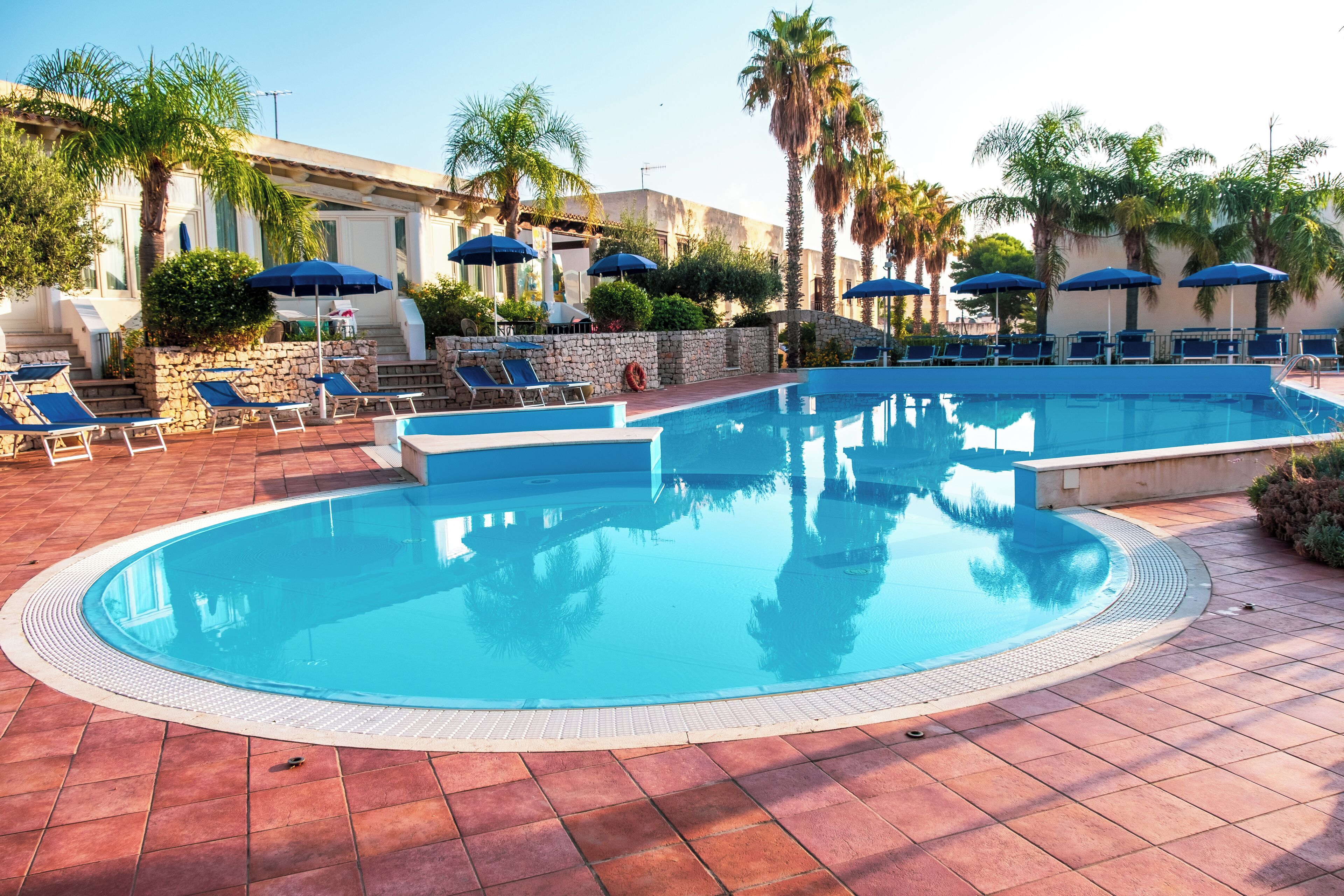 A pool area with lounge chairs and umbrellas, surrounded by palm trees and a stone wall.