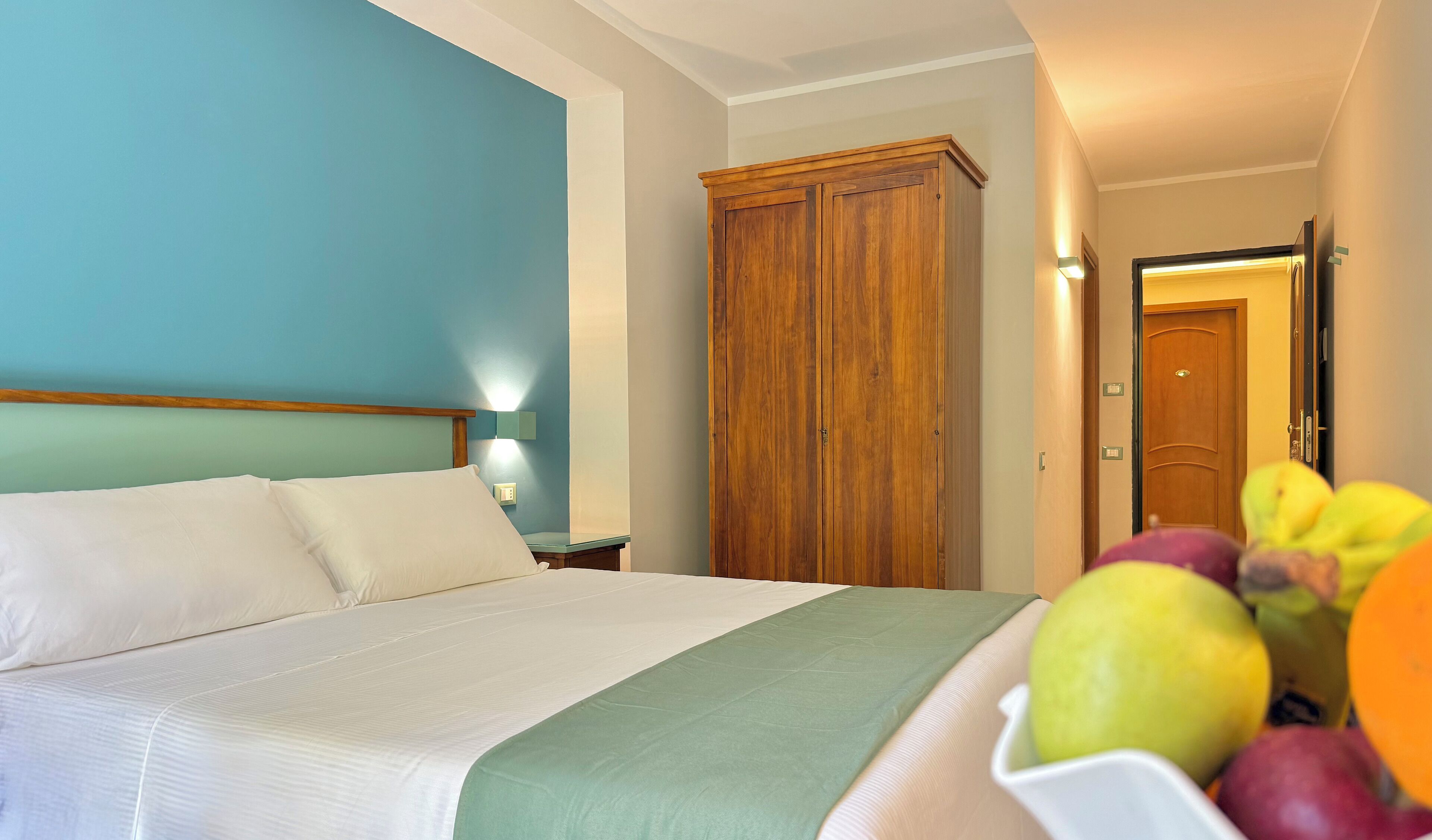 A hotel room with a bed, a wooden wardrobe, and a fruit bowl on the nightstand.
