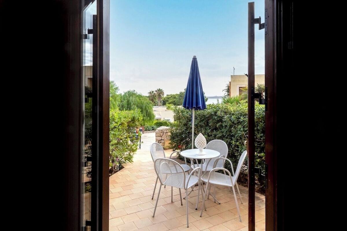 An outdoor patio with white metal chairs and a table, a blue umbrella, and a stone wall.