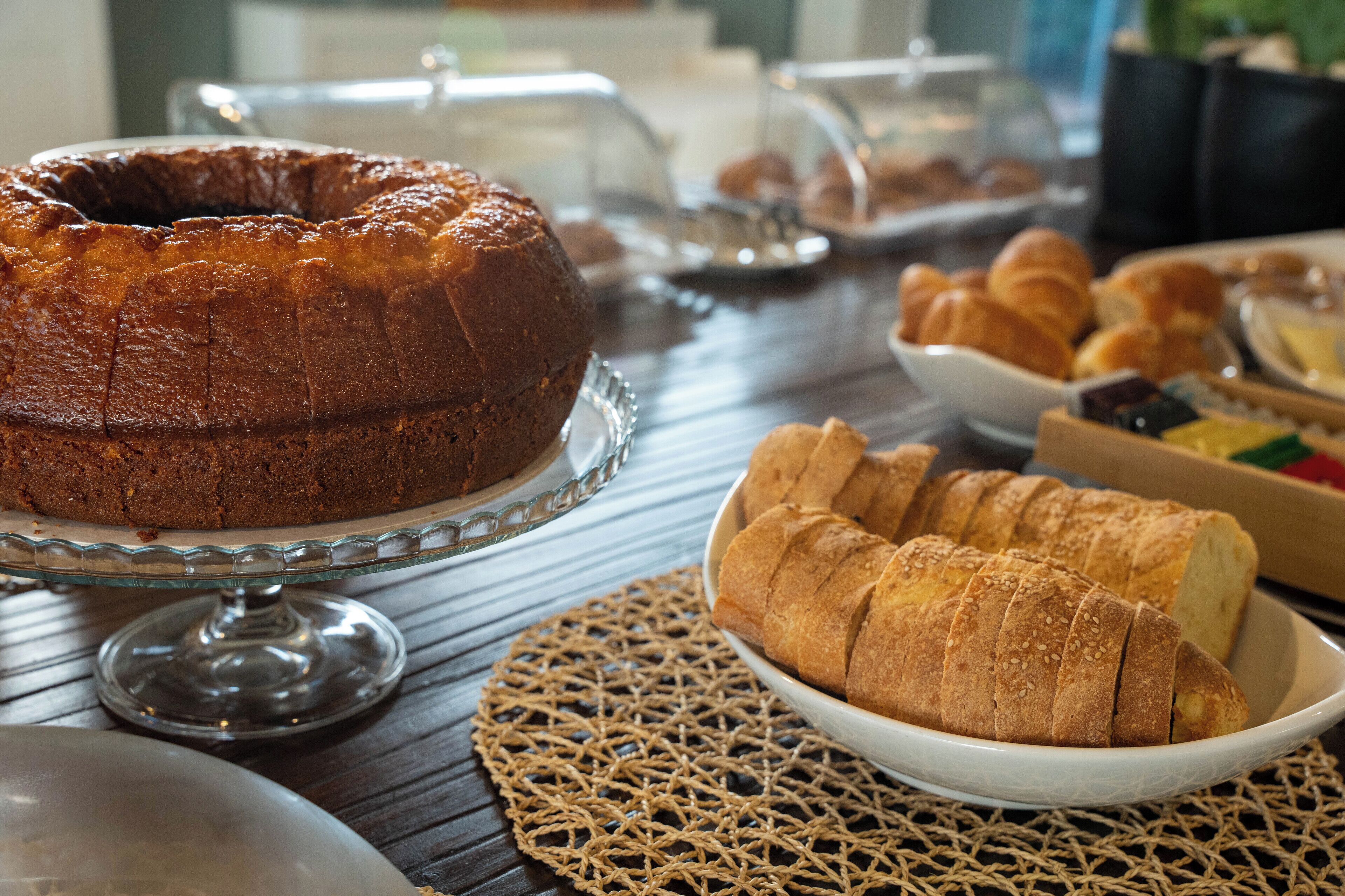 A wooden table with a cake stand holding a bundt cake, a plate with croissants, and a woven placemat.