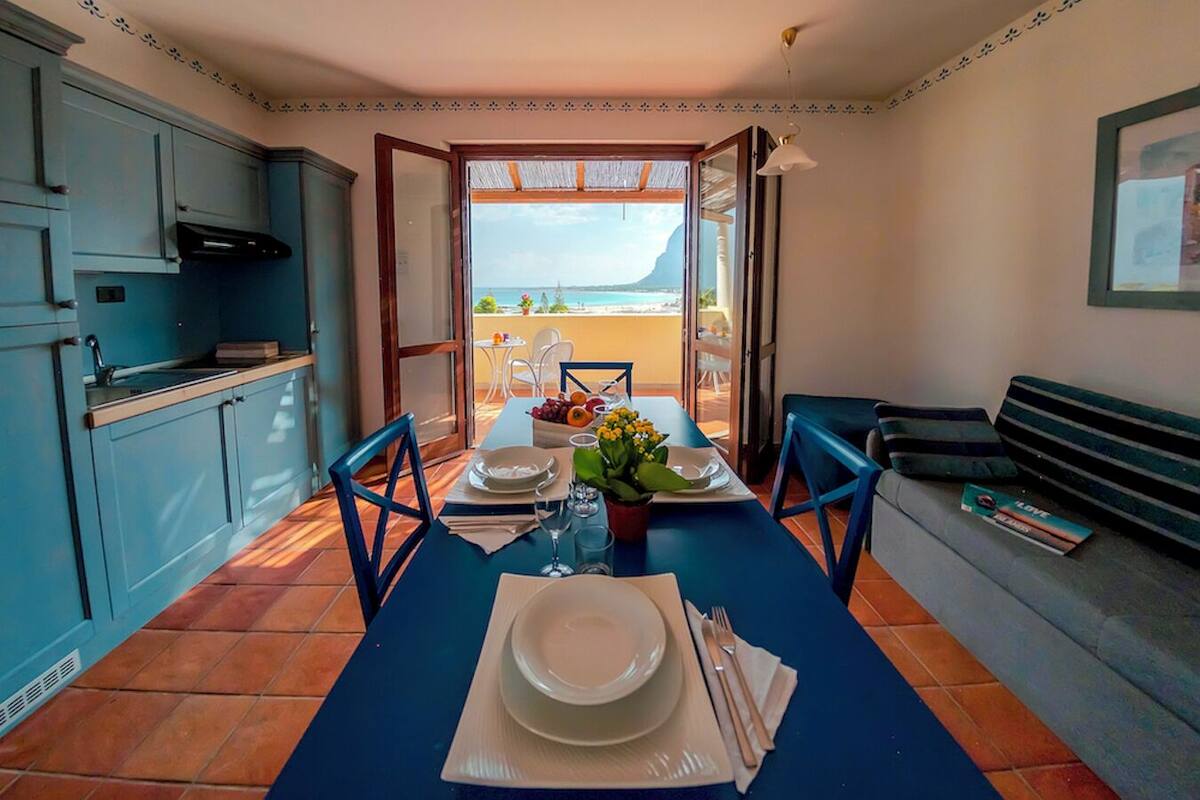 A dining area with a table set for two, a kitchen to the left, and a view of the sea through an open door.