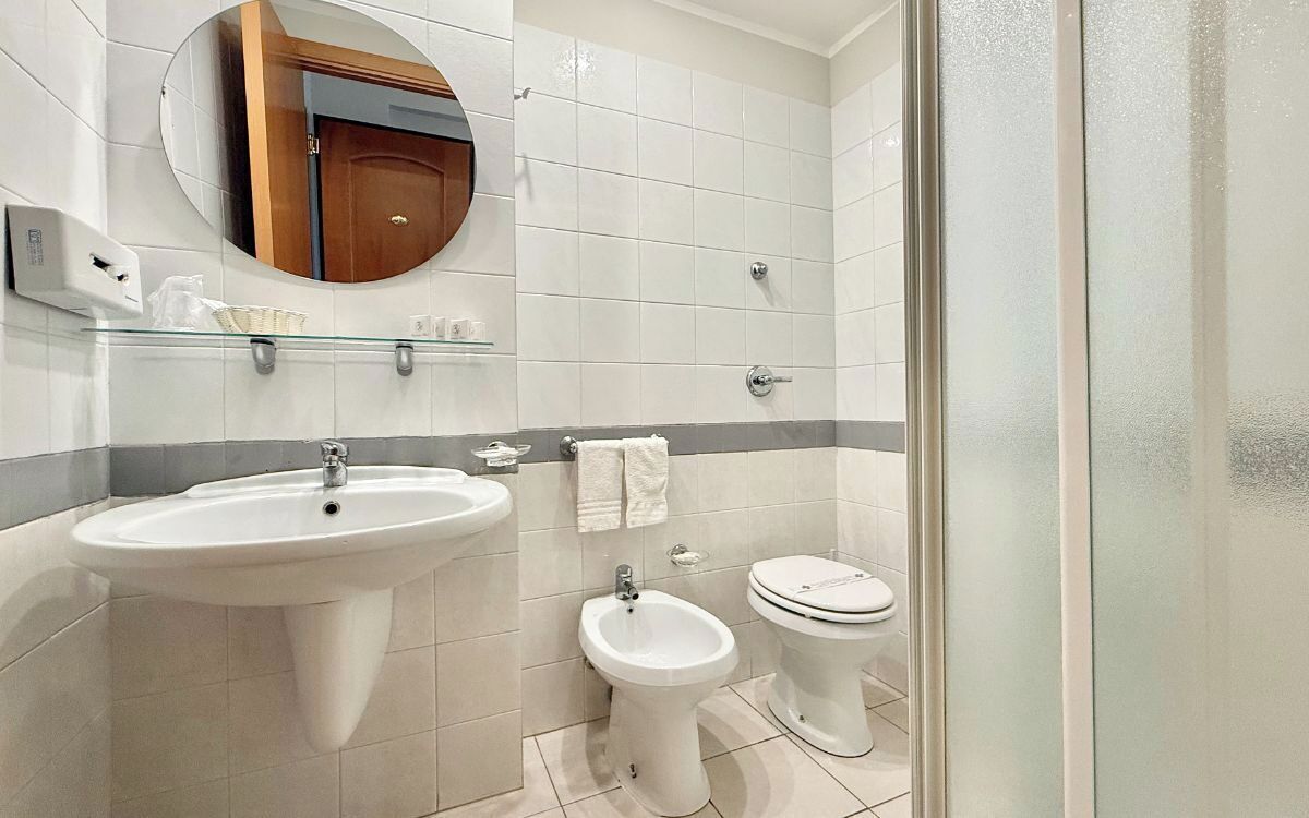 Un baño moderno con lavamanos pedestal, un bidé, un inodoro y un estante de vidrio sobre el lavamanos.