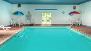 Una piscina interior con agua cristalina, paredes blancas y suelo de baldosas. Hay dos sombrillas, un cartel de advertencia y una puerta que da al exterior.