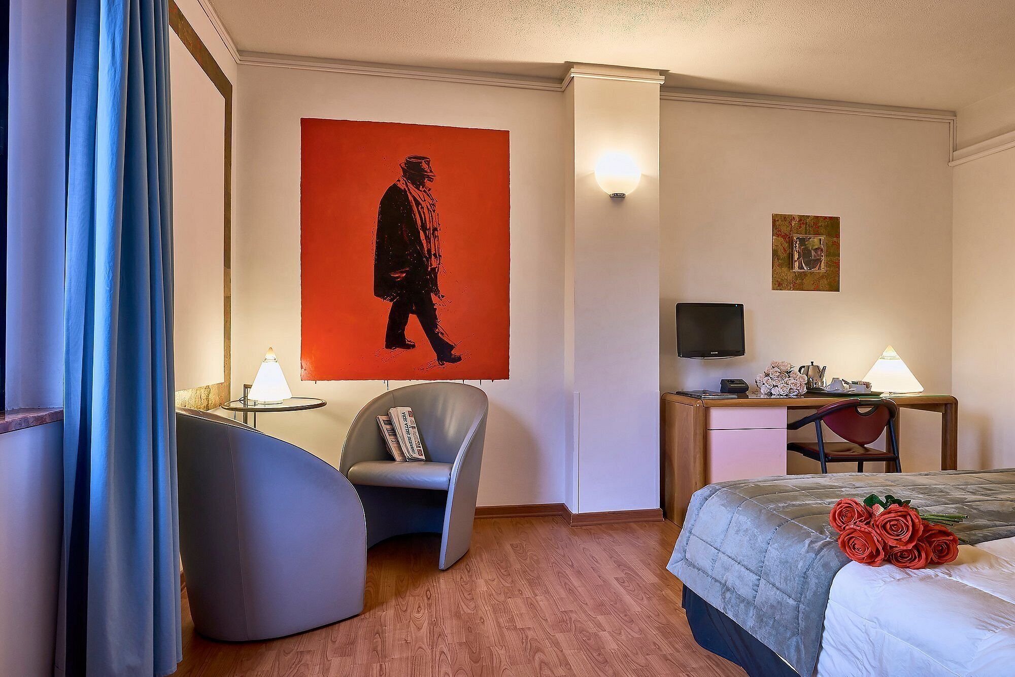 A hotel room with a bed, a desk, a chair, a wall-mounted TV, and a large painting.