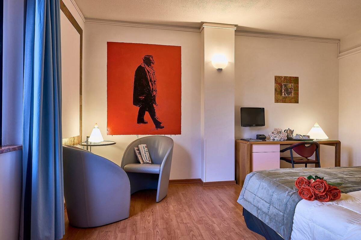 A hotel room with a bed, a desk, a chair, a wall-mounted TV, and a large painting.