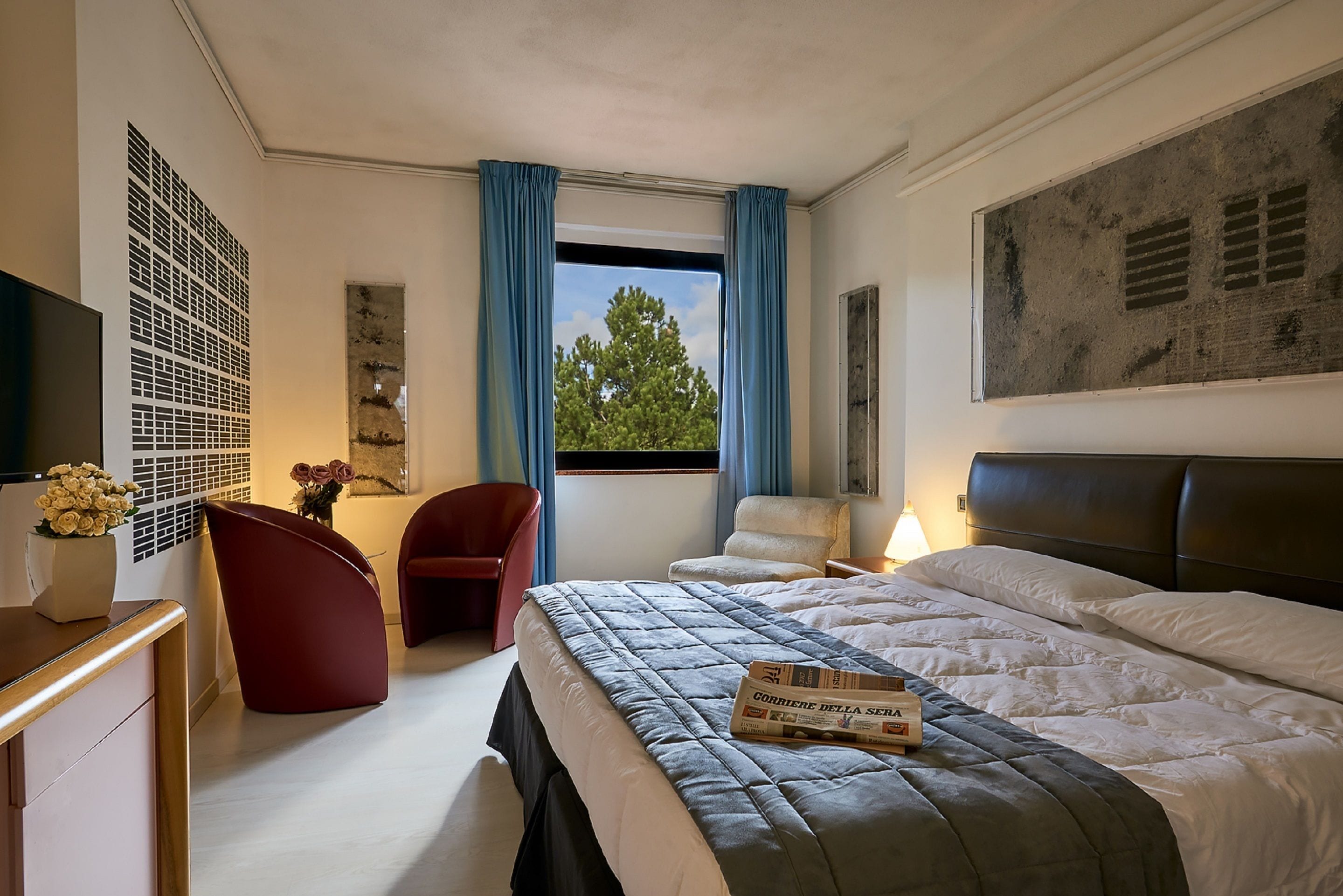 A modern hotel room with a large bed, a desk, a chair, and a window with curtains.