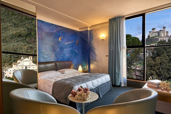 A hotel room with a large bed, two chairs, a small table with flowers, a nightstand, and a view of a castle through the window.