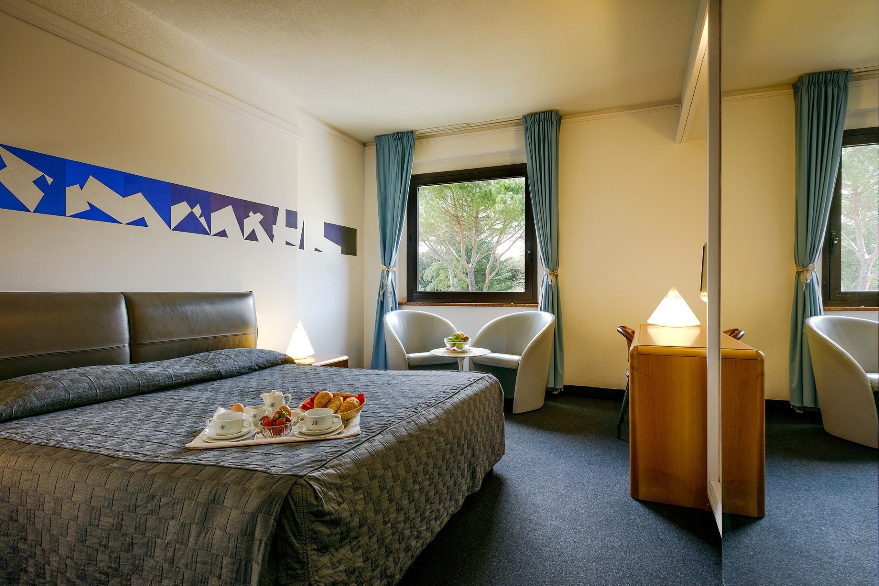 Een hotelkamer met een groot bed, een klein tafeltje met een lamp, twee stoelen en uitzicht op het landschap door een raam.