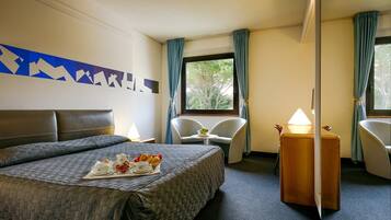 Een hotelkamer met een groot bed, een klein tafeltje met een lamp, twee stoelen en uitzicht op het landschap door een raam.