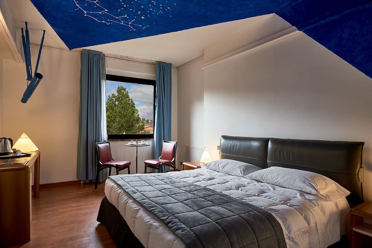 A hotel room with a bed, a desk with a lamp, a chair, and a view of the outdoors through a window.