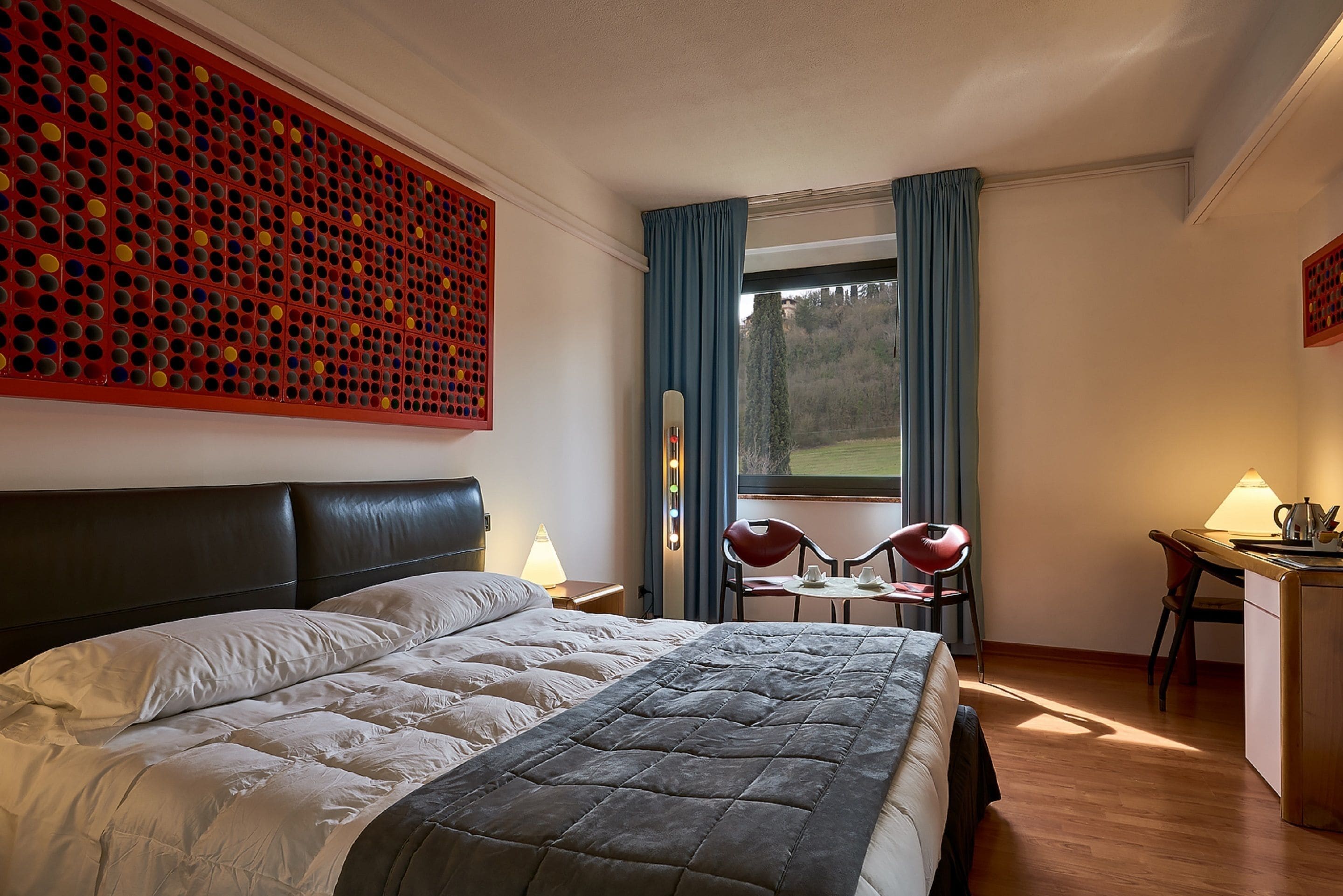 Habitación de hotel con una cama grande, un escritorio con una silla, una mesita y vistas al exterior a través de una ventana con cortinas.