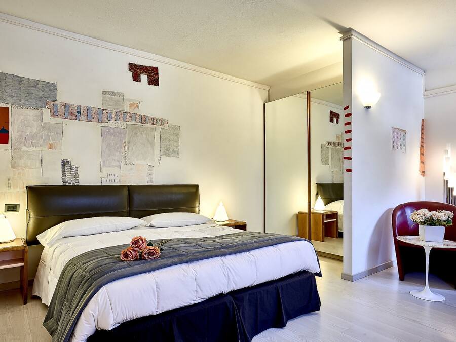 A hotel room with a large bed, a bedside table with a lamp, a chair with a flower pot, and a mirror on the wall.