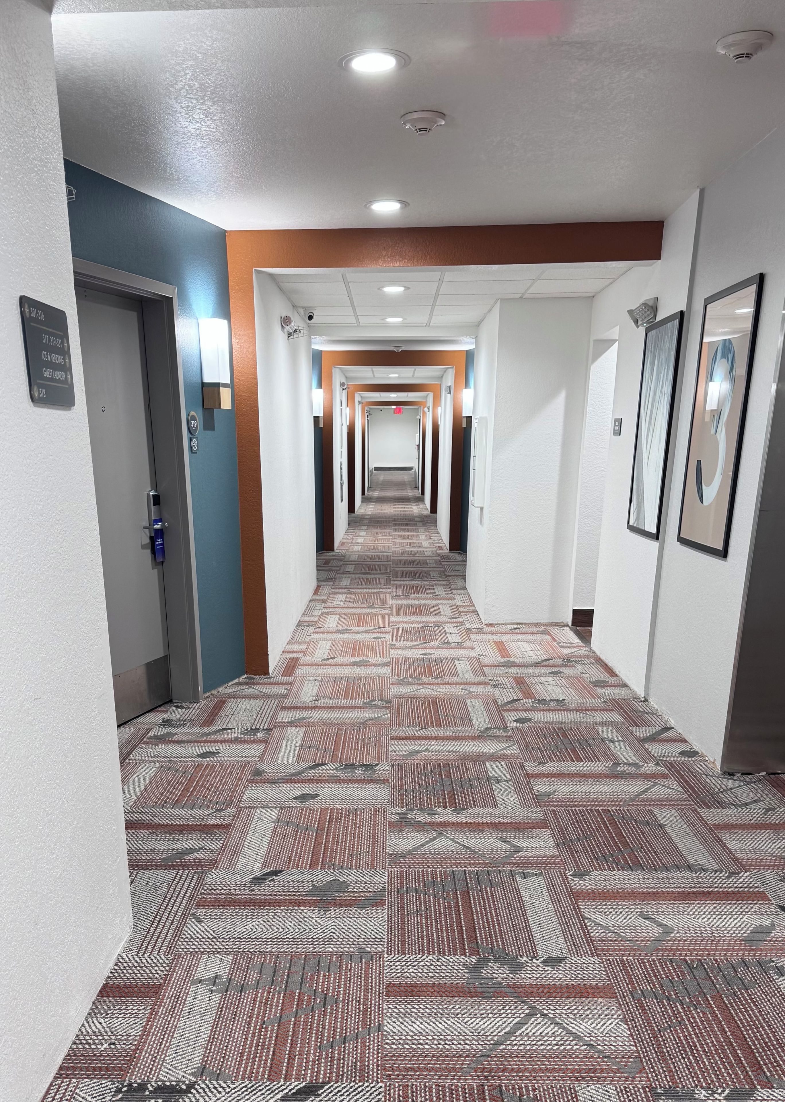 Un pasillo de hotel con alfombra estampada, paredes blancas y varias puertas.