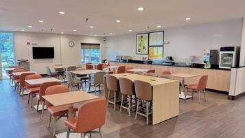 A modern dining area with tables and chairs, a kitchen counter, and a flat-screen TV on the wall.
