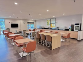 A modern dining area with tables and chairs, a kitchen counter, and a flat-screen TV on the wall.