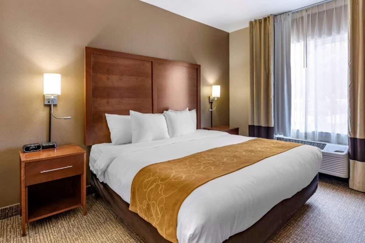 Una habitación de hotel con una cama grande, dos mesitas de noche, una lámpara de pared, un equipo de aire acondicionado y una ventana con cortinas.