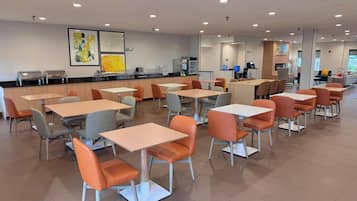 A modern cafeteria with multiple tables and chairs, a counter with food service equipment, and artwork on the walls.