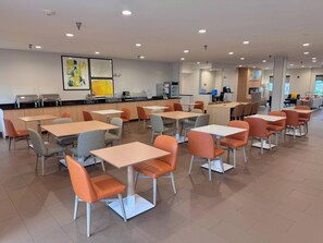 A modern cafeteria with multiple tables and chairs, a counter with food service equipment, and artwork on the walls. - Comfort Suites Coralville I-80 (Coralville)
