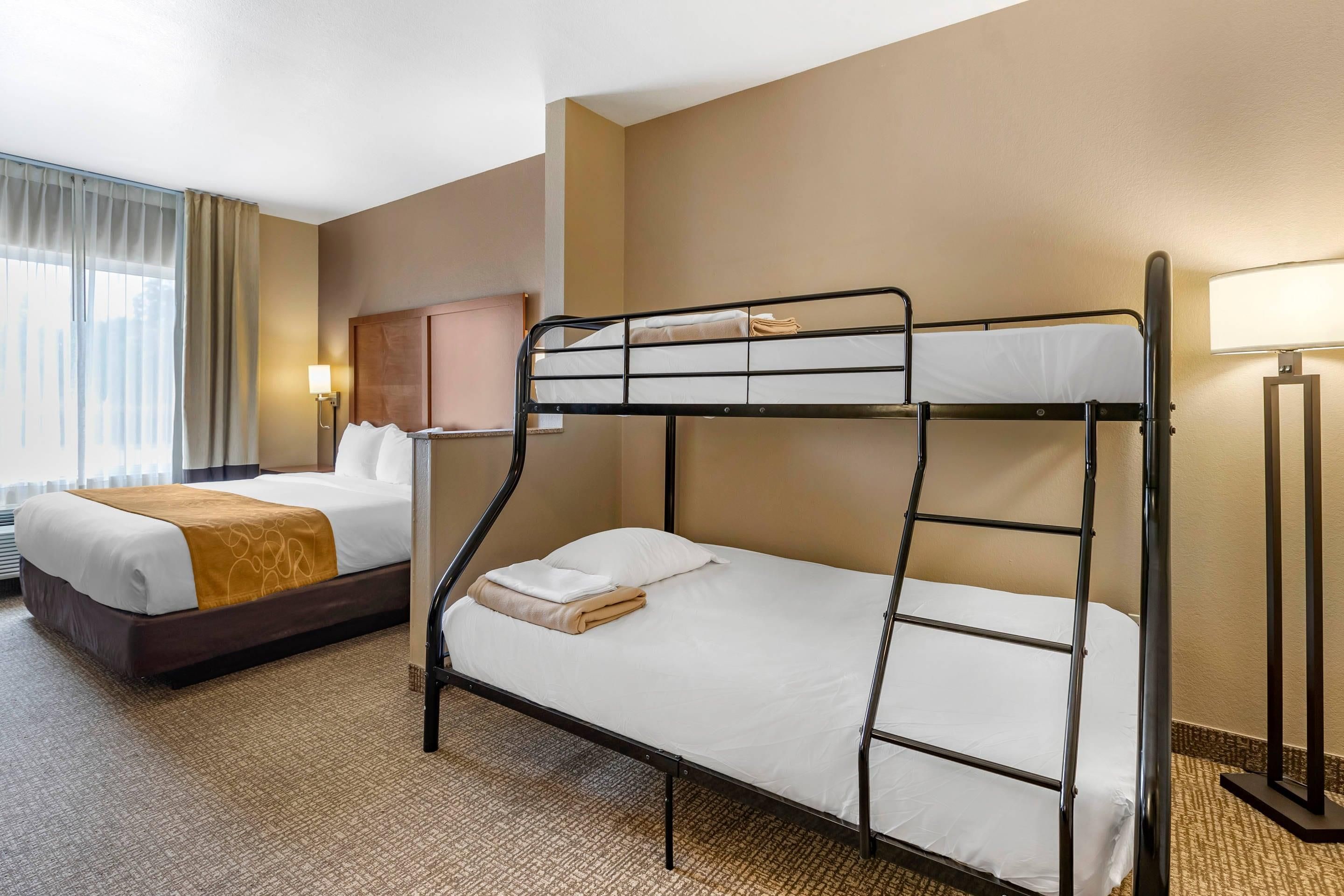 A hotel room with a bunk bed, a separate bed, a lamp, and a window with curtains.