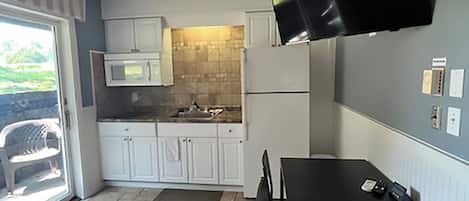 A compact kitchen with white cabinets, a microwave, a flat-screen TV mounted on the wall, a ceiling fan, and a dining table with chairs.