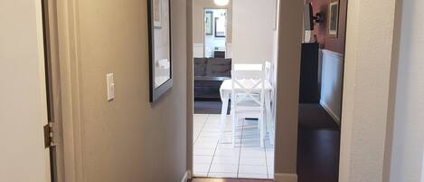 A hallway leading to a living area with a couch and chairs, a door on the left, and a radiator on the right.