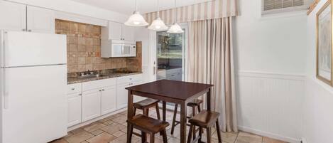 Una cocina con armarios blancos, una mesa de madera oscura y dos taburetes de bar. Hay una ventana con cortinas y un equipo de aire acondicionado en la pared.