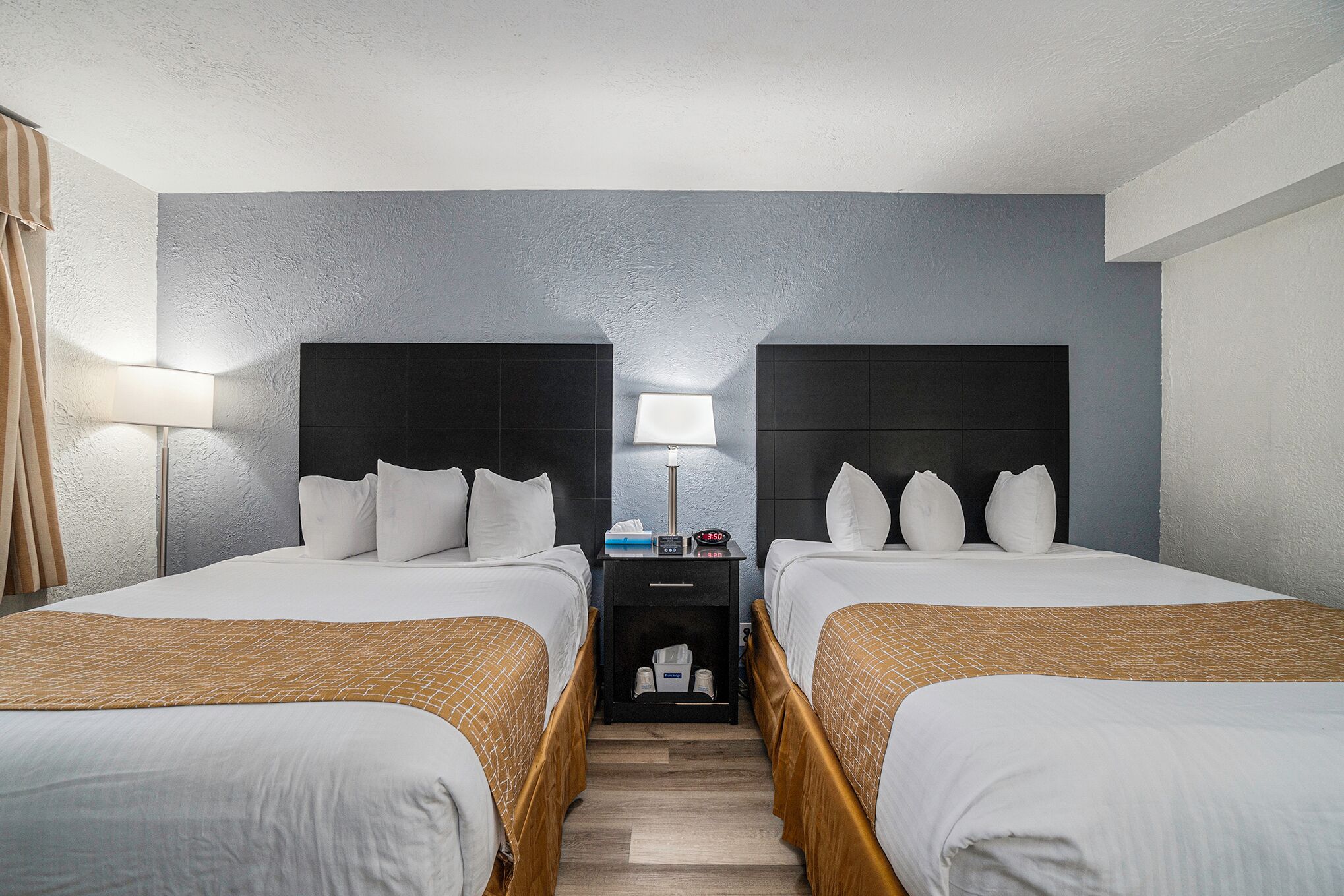 A hotel room with two beds, a nightstand with a lamp, and a small storage area.