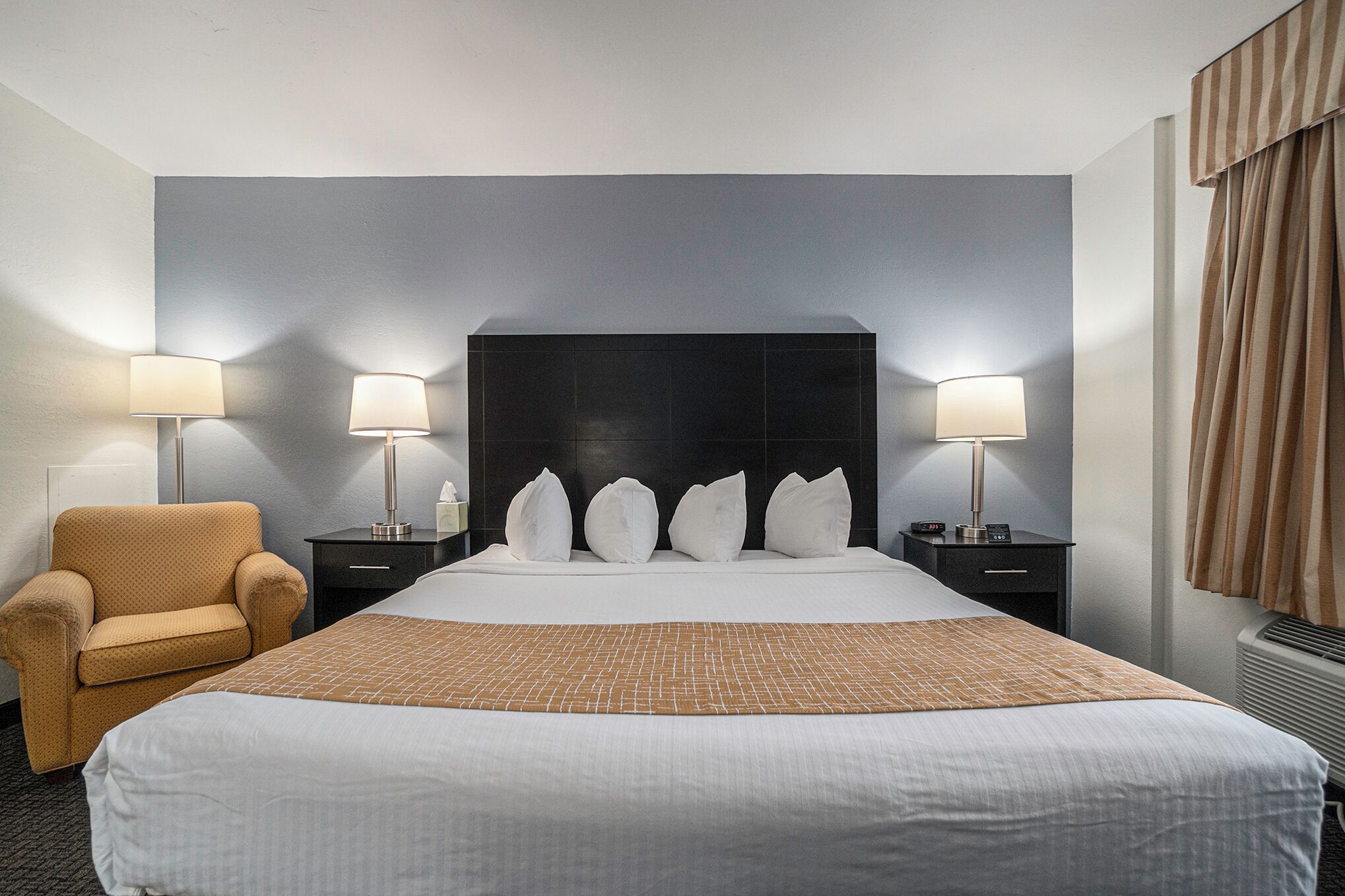 Une chambre d’hôtel avec un grand lit, deux lampes de chevet, une chaise et une tête de lit sombre.