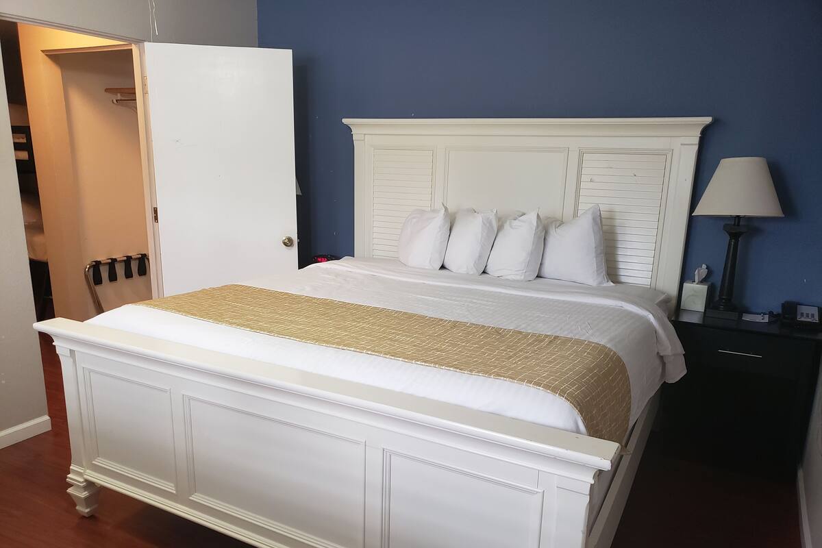 Un lit bien fait, avec une tête de lit blanche rehaussée d’une touche dorée, une table de chevet avec une lampe et une commode sombre.