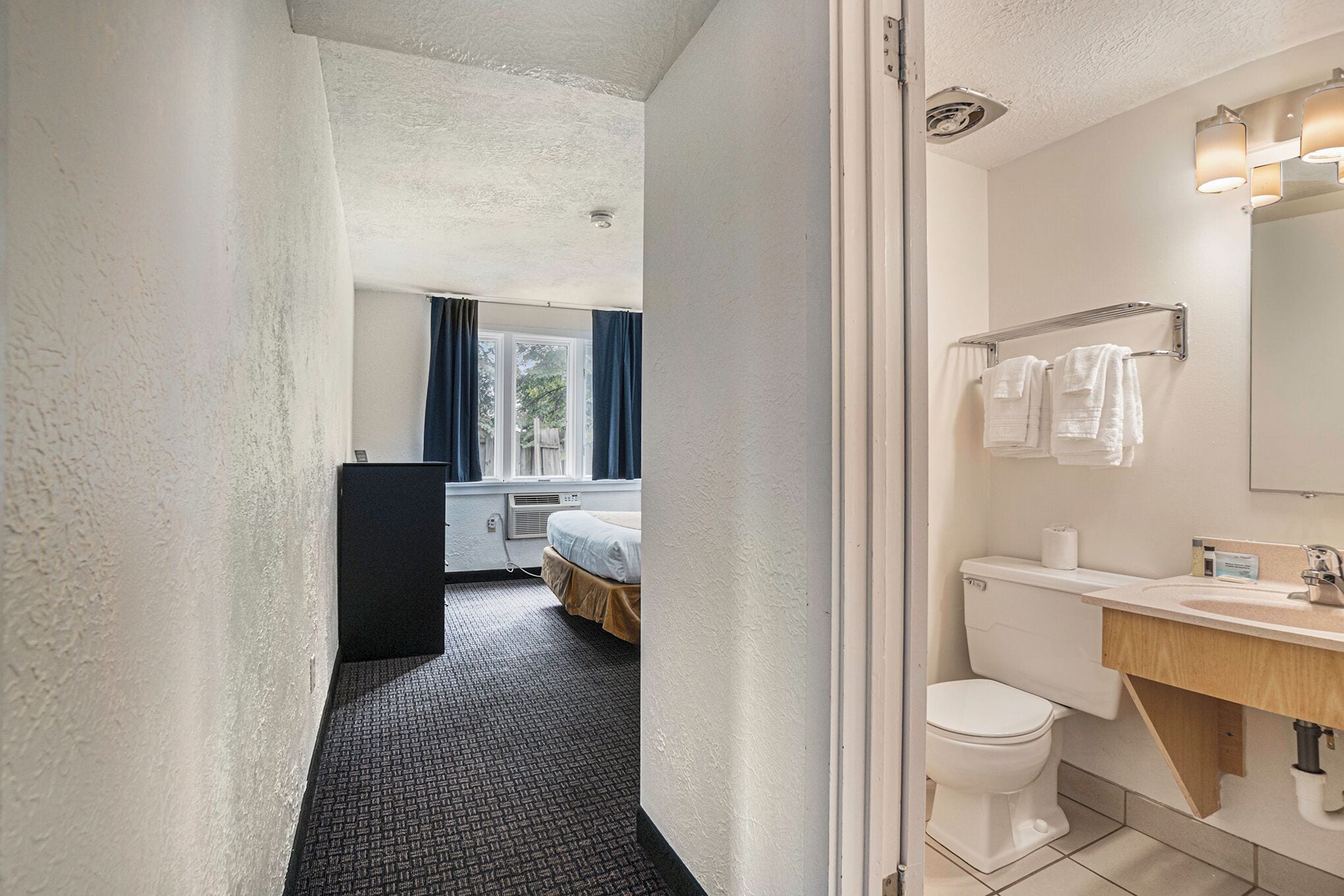 A hotel room with a bathroom featuring a toilet, sink, and mirror. There is a bed with a window and curtains in the room.