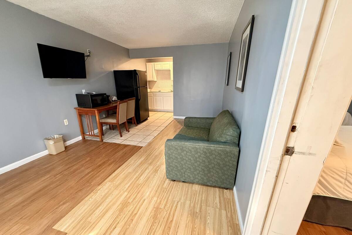 A living room with a wooden floor, a green sofa, a dining table with chairs, a black refrigerator, and a flat-screen TV mounted on the wall.