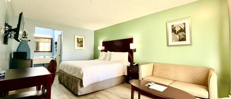 A hotel room with a bed, a sofa, a desk, and a chair. There is a window and a door visible.